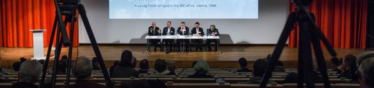 Ciemna widownia, oświetlona scena, przy stole prezydialnym naukowcy, którzy biorą udział w dyskusji, na ekranie za nimi czarno-białe zdjęcie przedstawia dziecko, kryjące się za dorosłymi