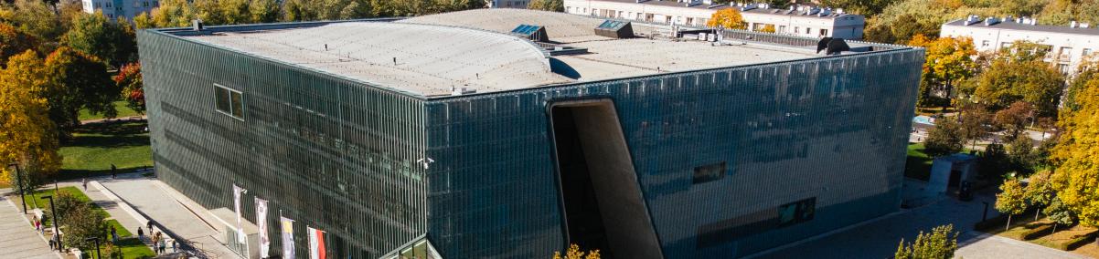 Budynek Muzeum POLIN. Dookoła drzewa. W tle widać bloki, a po prawej stronie zdjęcia jeden z wieżowców.