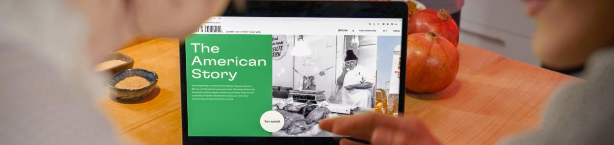 Zza pleców dwóch osób widać ekran laptopa, który stoi na blacie stołu. Na ekranie wyświetlona strona www "Od kuchni". Widać napis po angielsku: The American Story