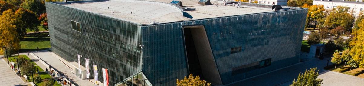 Budynek Muzeum POLIN prezentowany z zewnątrz. Wokół niego park, parking, plac i pobliskie bloki.