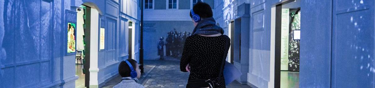 Kobieta z dzieckiem w słuchawkach spacerują po galerii "Na żydowskiej ulicy" na wystawie stałej w Muzeum POLIN.