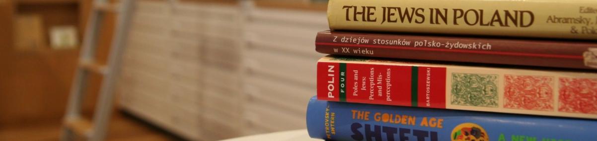 Grzbiety książek ułożonych jedna na drugiej. Leżą na blacie stołu. W głębi regały biblioteczne