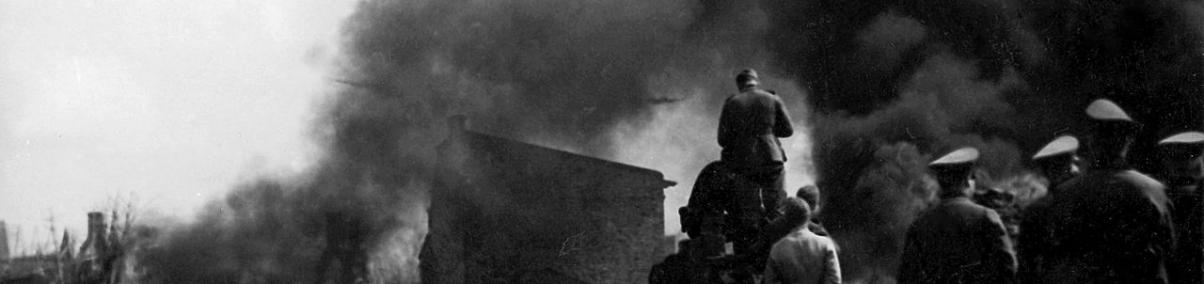 Kłęby dymu nad Warszawą w 1939 r. Na niebie leci samolot. Obserwuje to grupa kilku osób w mundurach i cywilów.