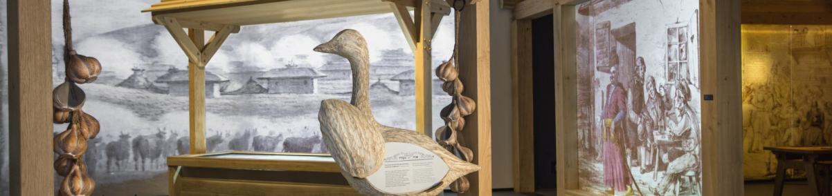 Galeria Miasteczko na wystawie Muzeum POLIN - dość ciemna przestrzeń, w której stoi stragan, na nim drewniana gęś i wianki drewnianych główek czosnków. W tle ekrany wyświetlające ryciny ze scenami z życia Żydów.