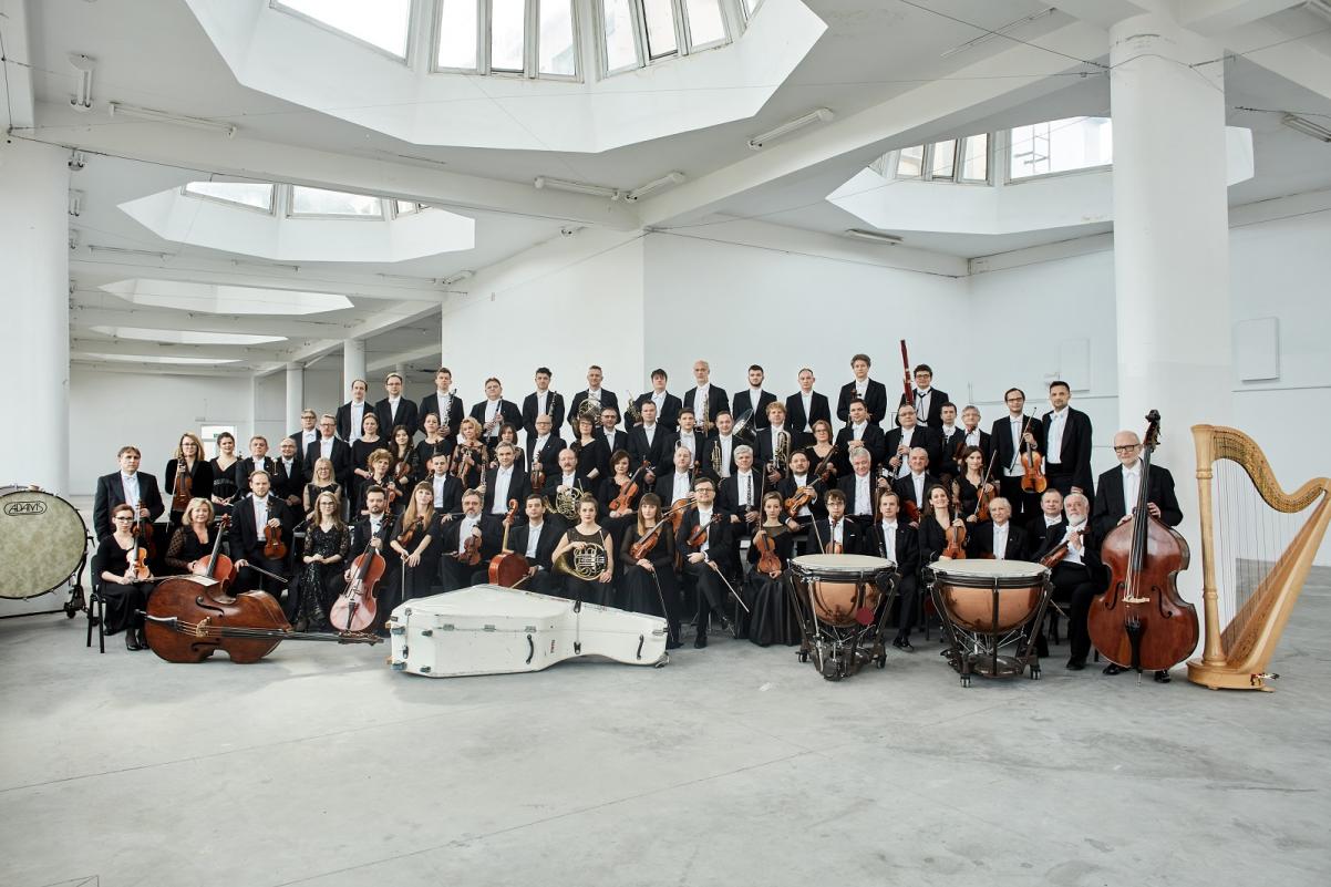 Na zdjęciu widać muzyków Orkiestry Sinfonia Varsovia w strojach koncertowych, wraz z instrumentami. Muzycy znajdują się w dużej jakby fabrycznej przestrzeni, z wieloma ogromnymi świetlikami w suficie