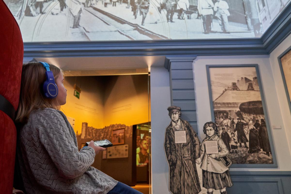 Z lewej strony zdjęcia dziecko, które siedzi na kanapie. W ręku trzyma audioprzewodnik, na uszach ma słuchawki. Patrzy na wystawę w Muzeum POLIN