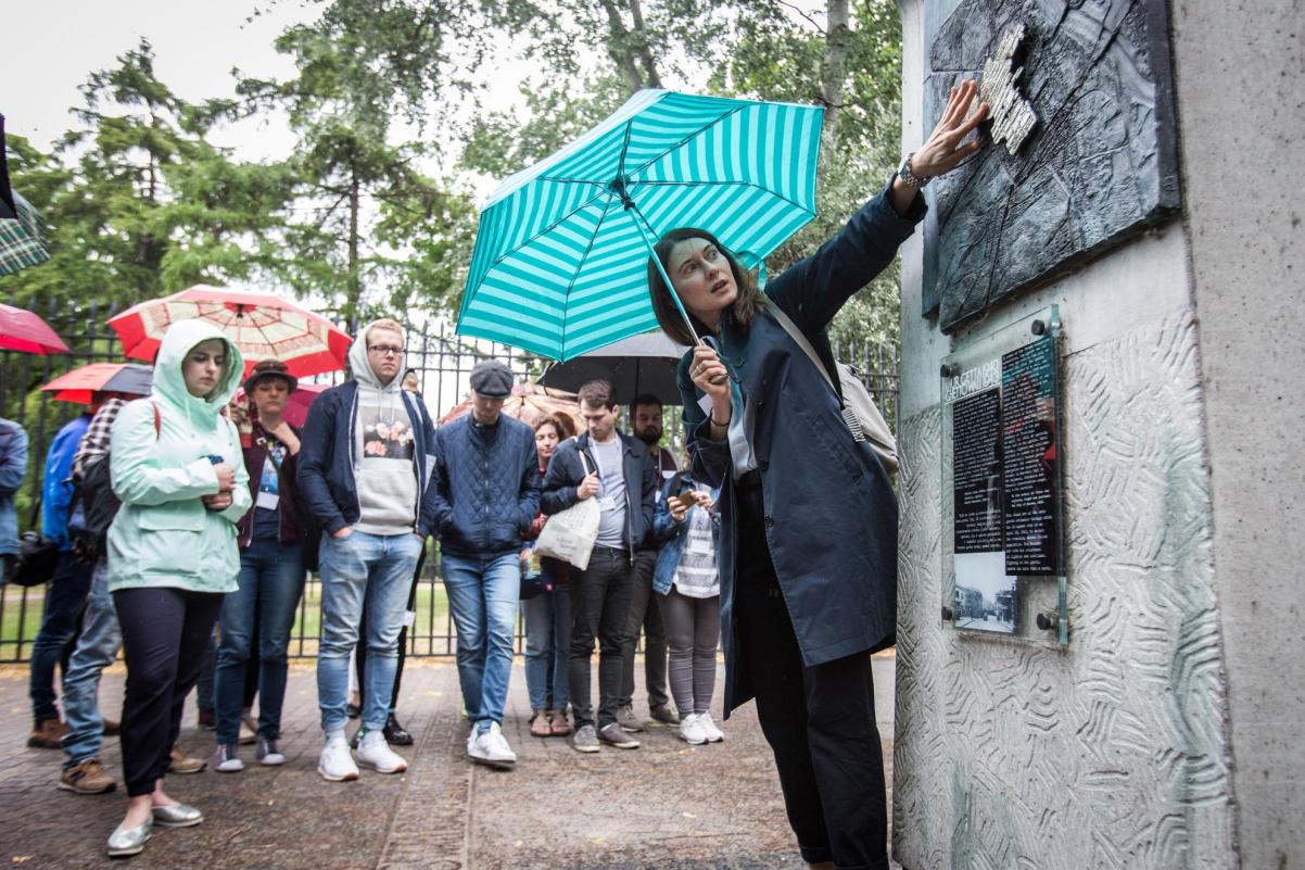 Na zdjęciu kobieta (przewodniczka) pokazuje element architektury i omawia go grupie zwiedzającej młodzieży. Deszczowy dzień, ludzie w kurtkach przeciwdeszczowych, pod parasolami