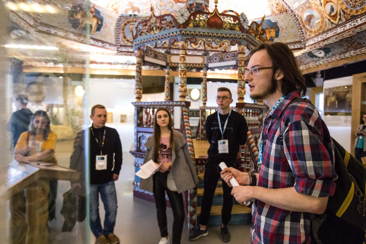 Grupa młodzieży znajduje się pod dachem zrekonstruowanej synagogi z Gwoźdźca, na wystawie stałej Muzeum POLIN. Sufit pięknie ozdobiony. Młodzież przygląda się wystawionym w gablocie zabytkom.