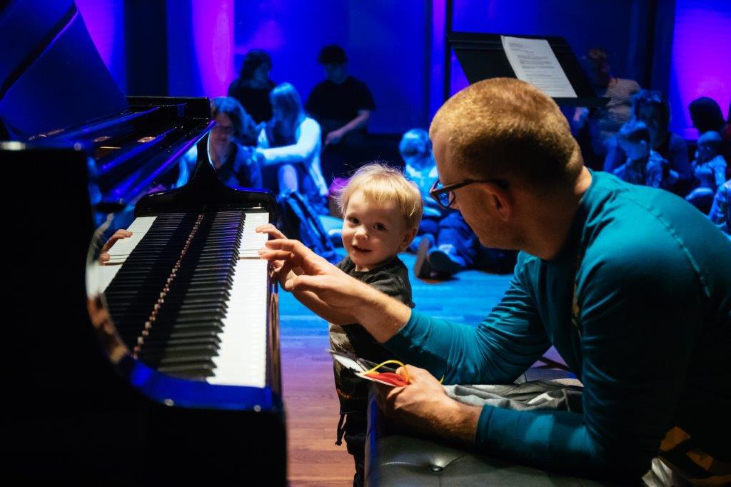 Przyciemniona przestrzeń. Mężczyzna pokazuje dziecku klawiaturę fortepianu. Dziecko się uśmiecha