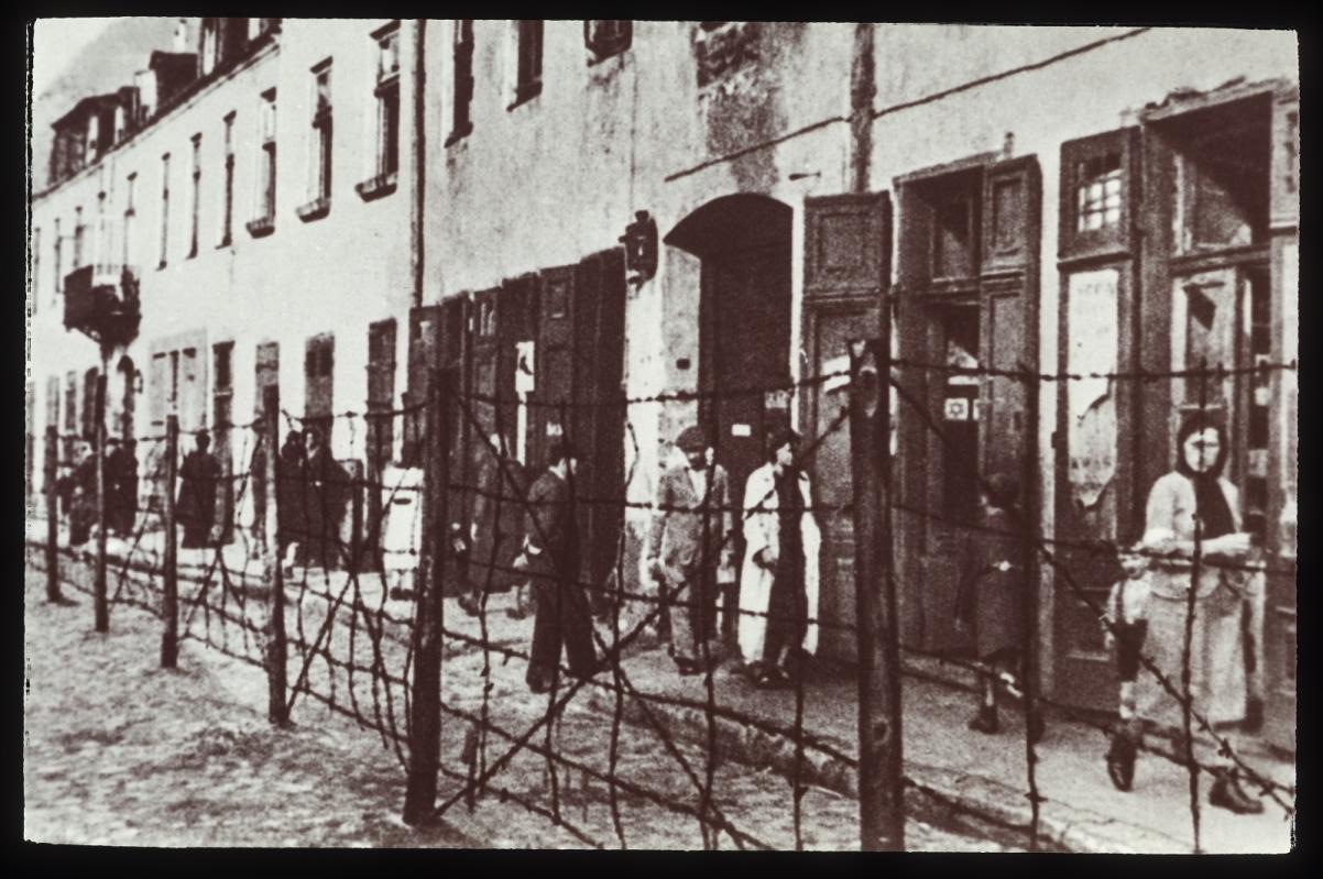 Archiwalne, czarno-białe zdjęcie getta warszawskiego, widziane zza ogrodzenia z drutu kolczastego. Zdjęcie znajduje się w zbiorach Muzeum POLIN
