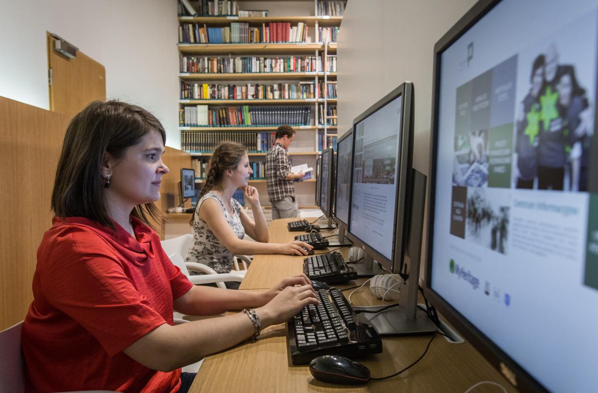 Młoda kobieta w czerwonej bluzce pracuje przy komputerze. W tle widać kolejne stanowiska komputerowe i księgozbiór.