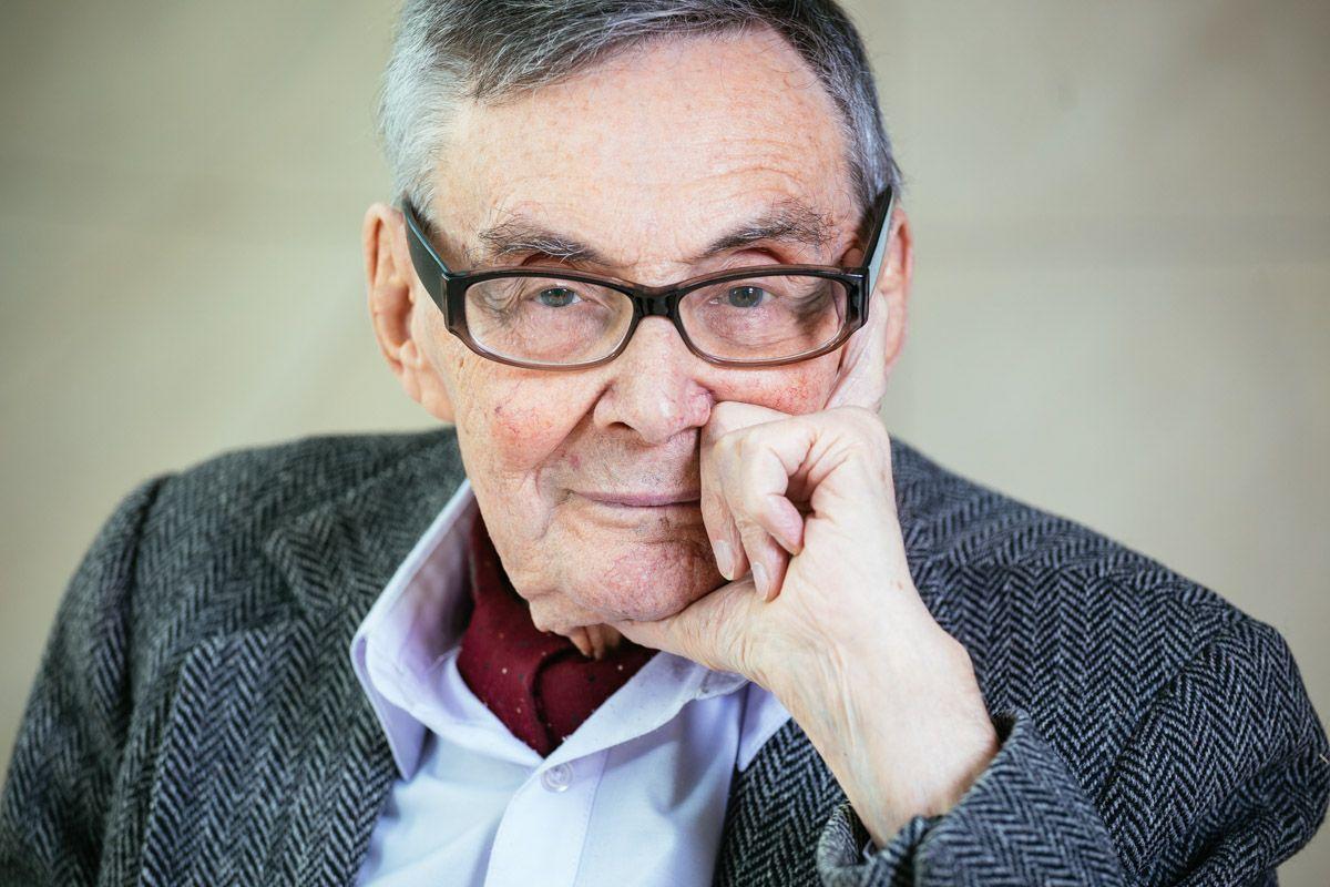 Marian Turski ubrany w niebieską koszulę i marynarkę. Podpiera twarz dłonią i patrzy przed siebie.