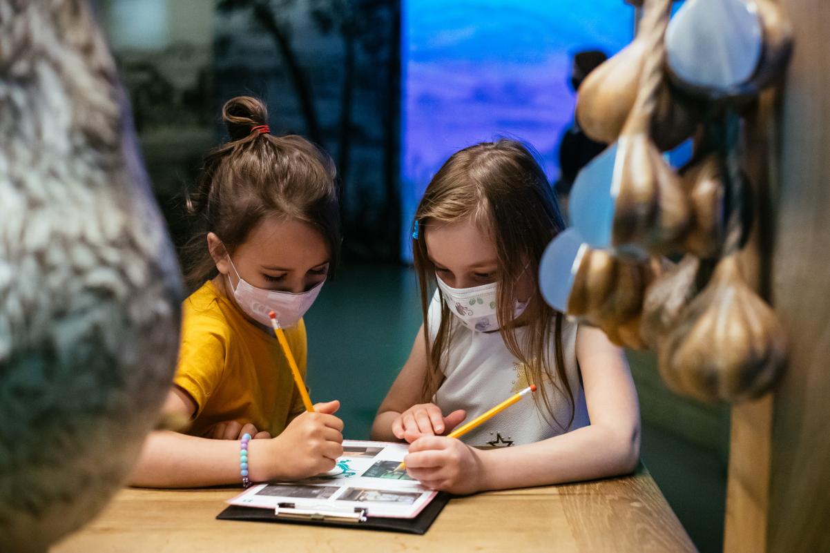 Dwoje dzieci w maseczkach notuje na kartce. Znajdują się przy straganie w galerii Miasteczko, na wystawie stałej Muzeum POLIN