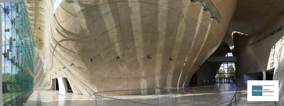 Hol główny Muzeum POLIN - widoczne wejście, ściana w kolorze piaskowym i ogromne niebieskawe okno po lewej stronie. W prawym dolnym rogu logotyp Centrum Konferencyjnego Muzeum POLIN