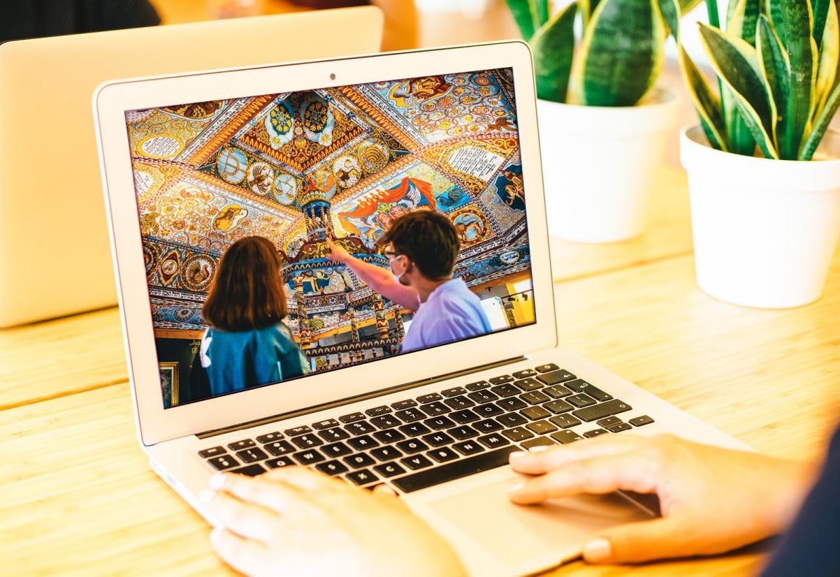 Na ekranie laptopa wyświetla się obraz dwojga zwiedzających oglądających synagogę na wystawie stałej POLIN. 