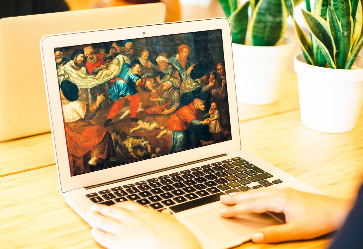 Otwarty laptop. Na ekranie wyświetla się obraz. W tle rośliny doniczkowe.