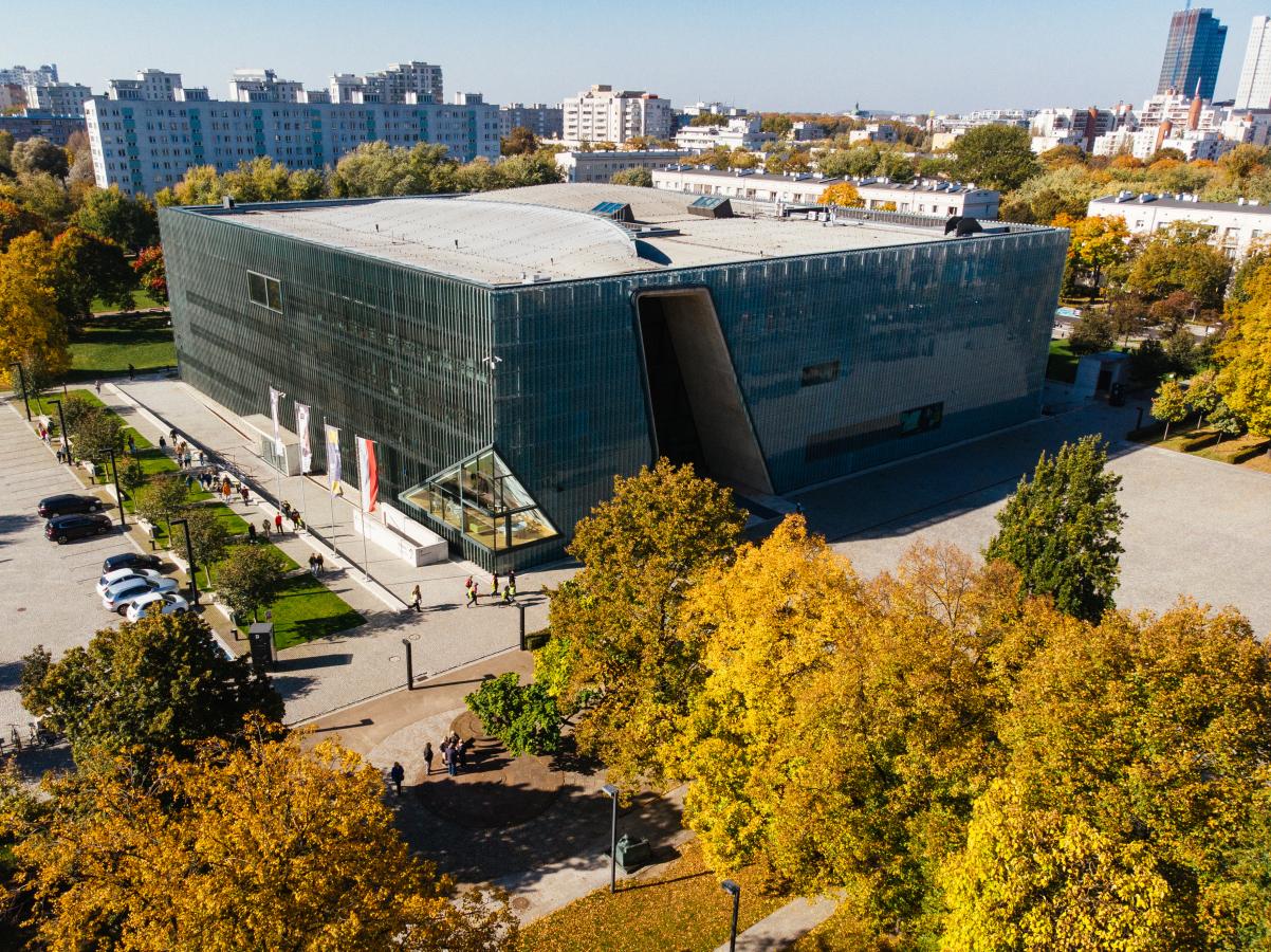 Budynek Muzeum POLIN. Dookoła drzewa. W tle widać bloki, a po prawej stronie zdjęcia jeden z wieżowców.