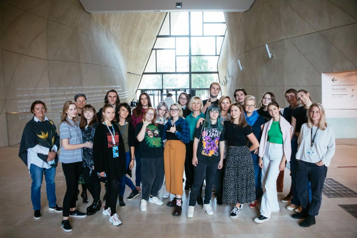 Duża grupa młodych osób stoi na korytarzy Muzeum POLIN, tyłem do głównego wejścia. Usmiechają się.