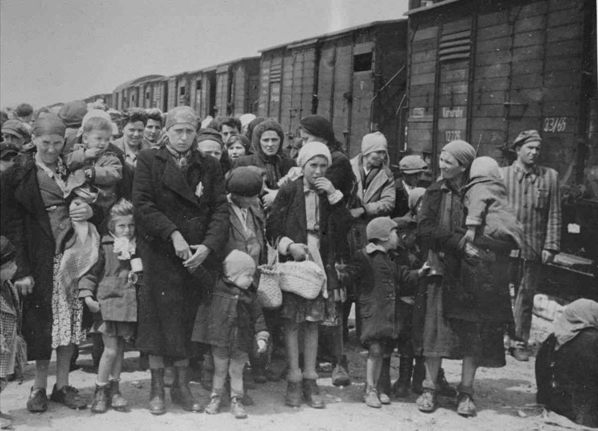 Grupa Żydów w różnym wieku stoi przy wagonach bydlęcych zmierzających do obozu zagłady.