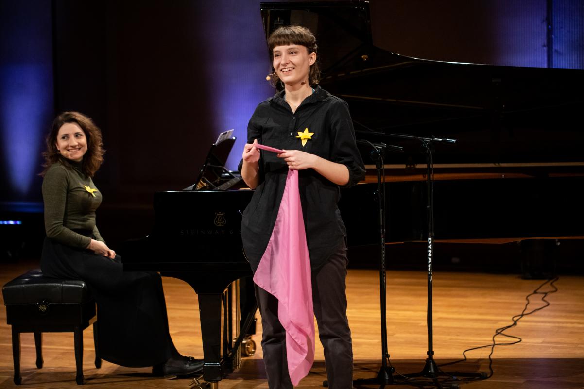 Na scenie znajdują się dwie kobiety w czarnych strojach. Jedna siedzi przy fortepianie, druga stoi i trzyma różową chustę.