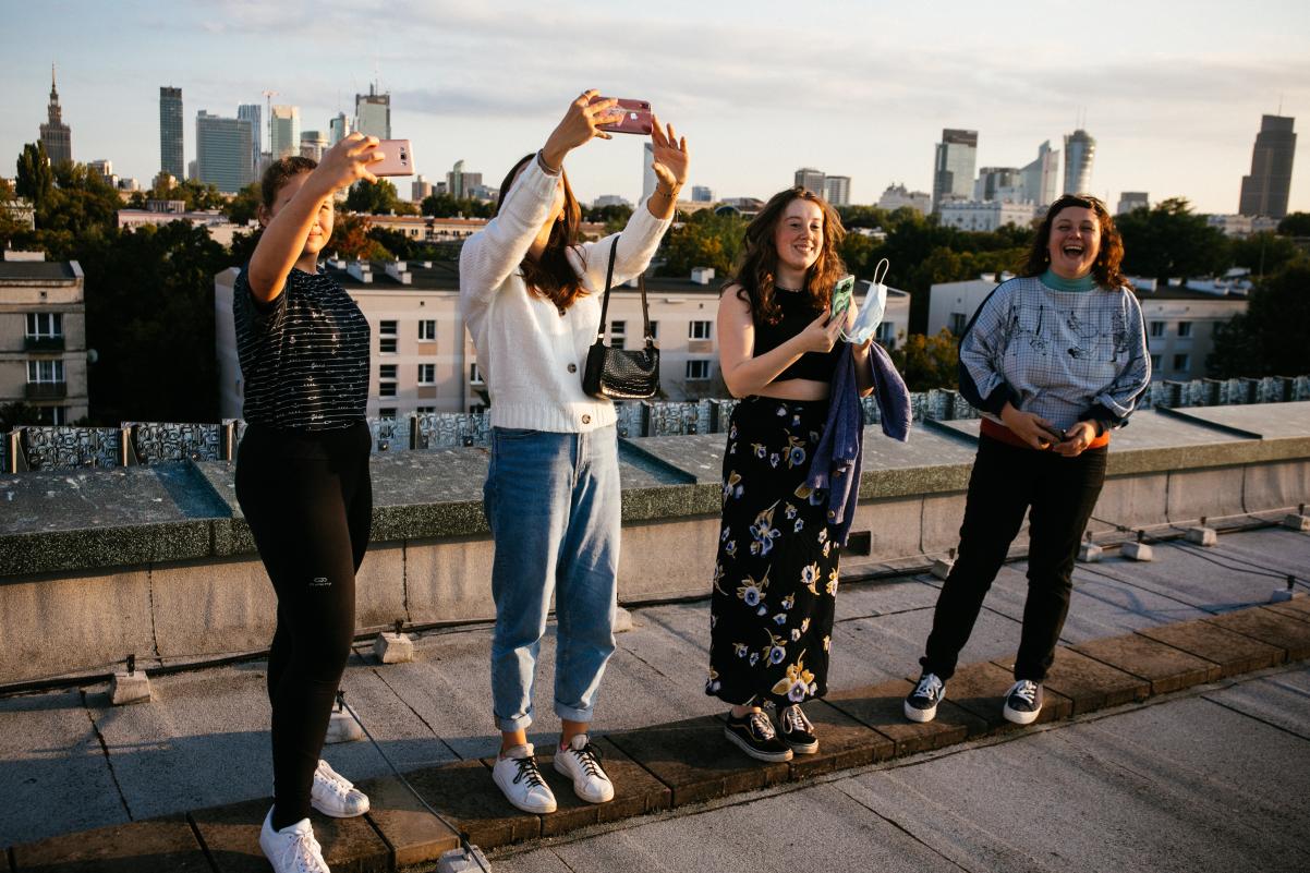 Na dachu budynku stoją rzędem cztery osoby w letnich ubraniach. Każda trzyma przed sobą telefon lub aparat fotograficzny. Robią zdjęcia. W dole za nimi panorama miasta.
