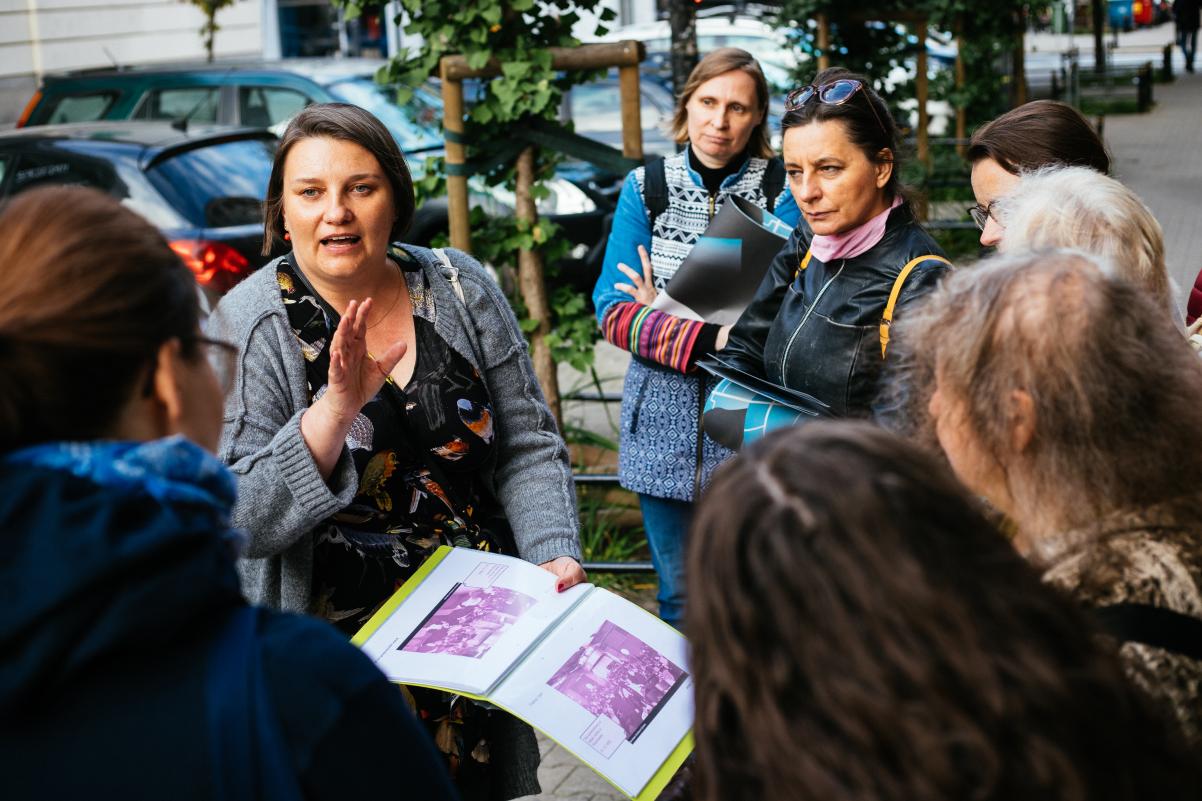Agnieszka Kuś trzyma w ręku graiki z planem Muranowa i opowiada zawartą w nich historię zgromadzonym wokół niej uczestnikom spaceru