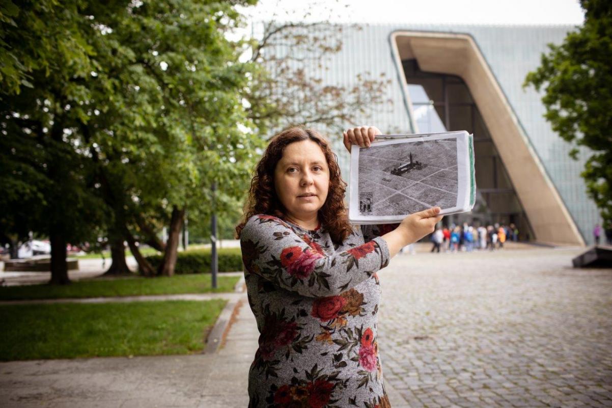 Przewodniczka Anita Borkowska stoi przed budynkiem Muzeum POLIN i trzyma zdjęcie getta warszawskiego. 
