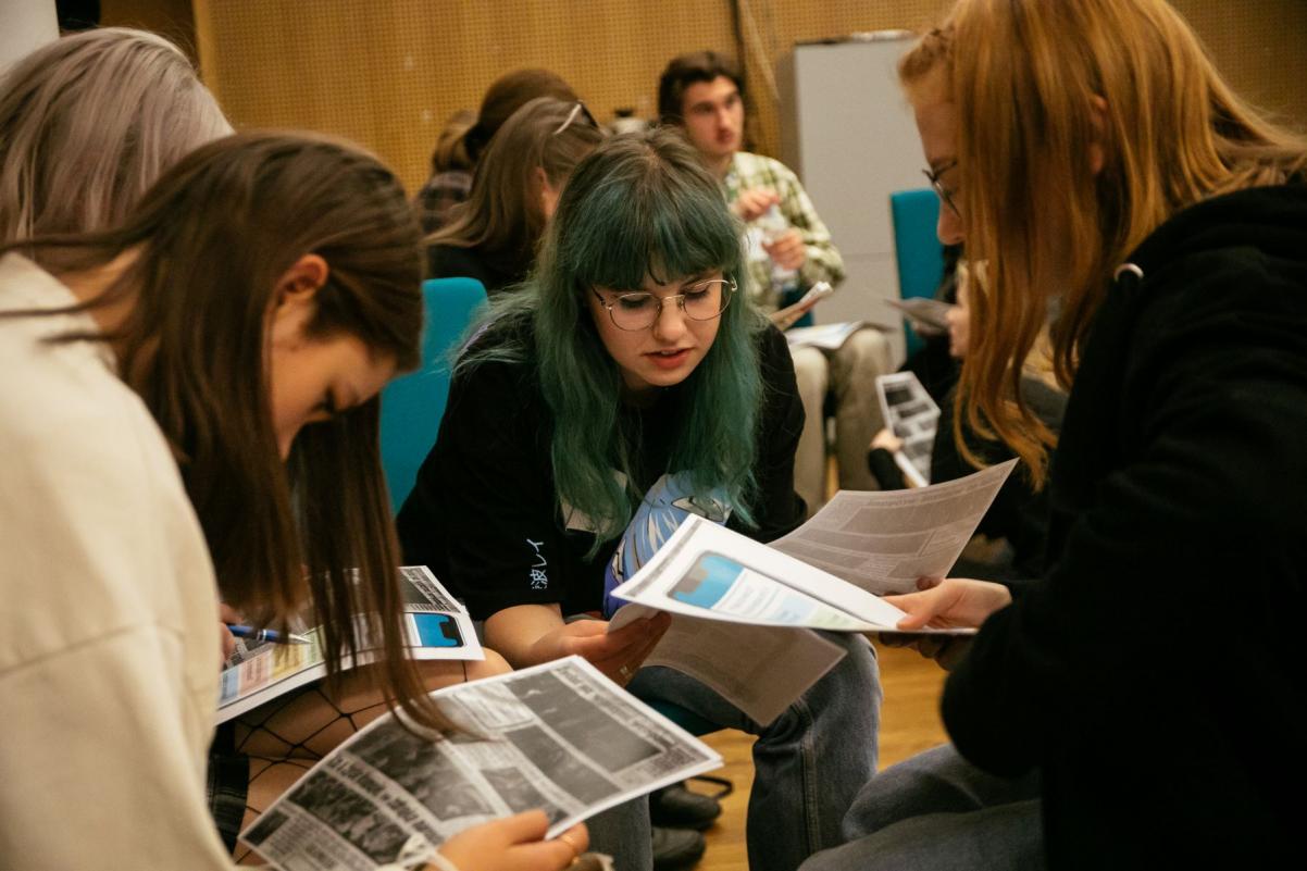 Cztery dziewczyny siedzą razem i czytają coś z kartek, które trzymają przed sobą.