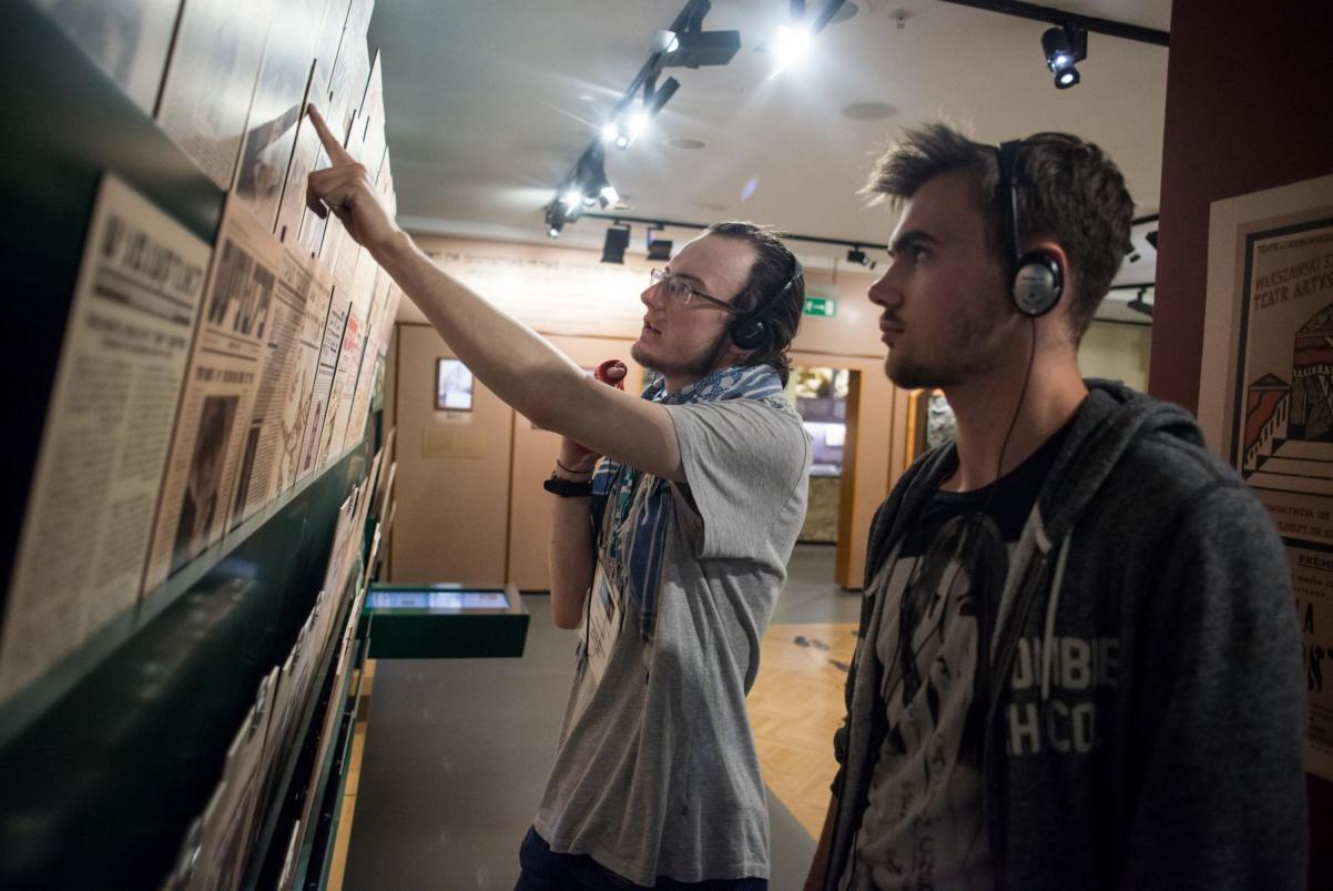 Dwaj młodzi mężczyźni oglądają gazety prezentowane na wystawie stałej w Muzeum POLIN. Jeden dotyka palcem gazetę.