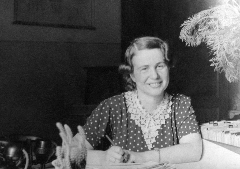 Irena Sendlerowa w biurze Wydziału Opieki Społecznej w Warszawie, lipiec 1949. Siedzi uśmiechnięta przy biurku, trzymając wieczne pióro.