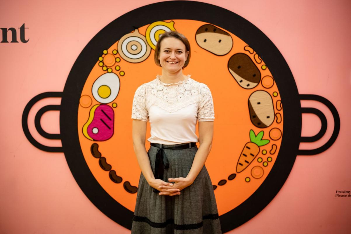 Tatsiana Kazak stoi na tle garnka z czulentem narysowanego na ścianie na wystawie "Od kuchni".