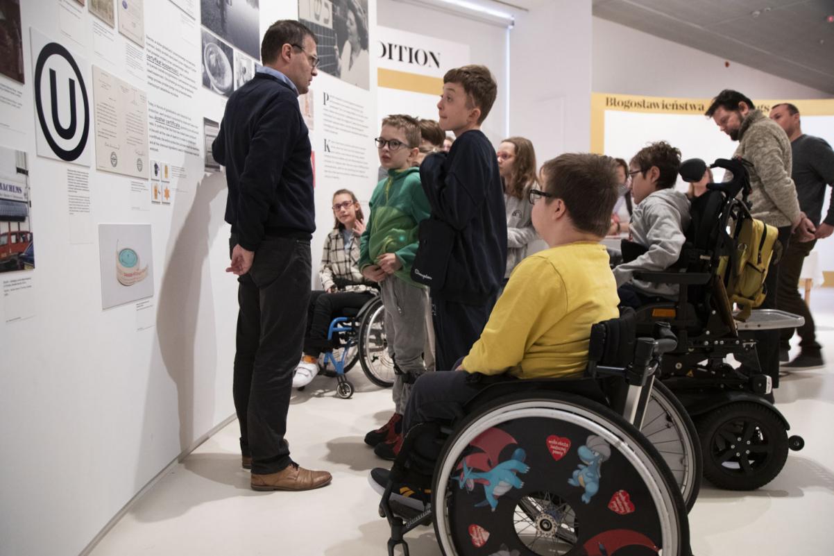 Dzieci z niepełnosprawnościami, w tym na wózkach inwalidzkich, zwiedzają wystawę "Od kuchni" z przewodnikiem.