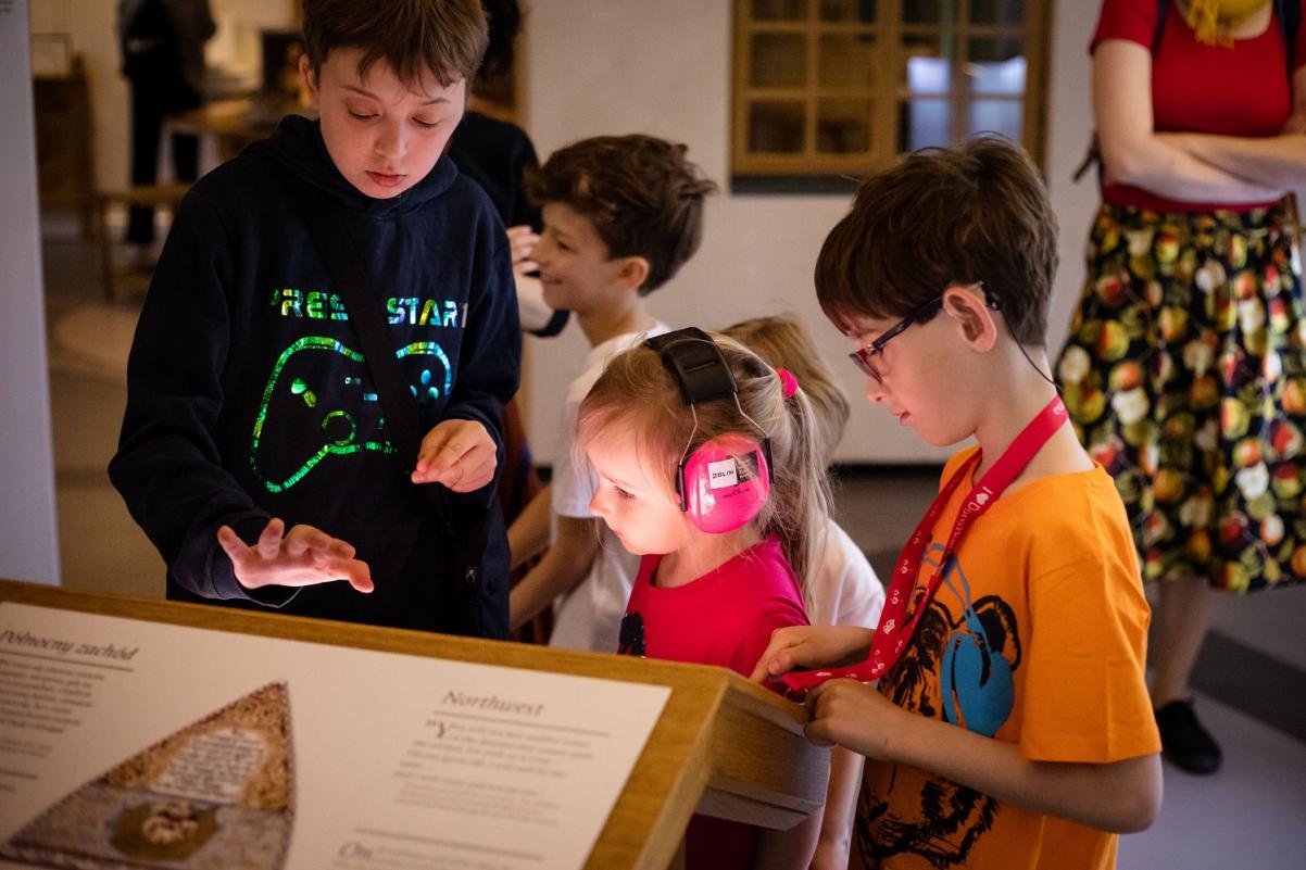 Troje dzieci stoi przy pulpicie z informacjami na wystawie w Muzeum POLIN. Między dwoma chłopcami znajduje się dziewczynka, która ma na uszach różowe słuchawki.