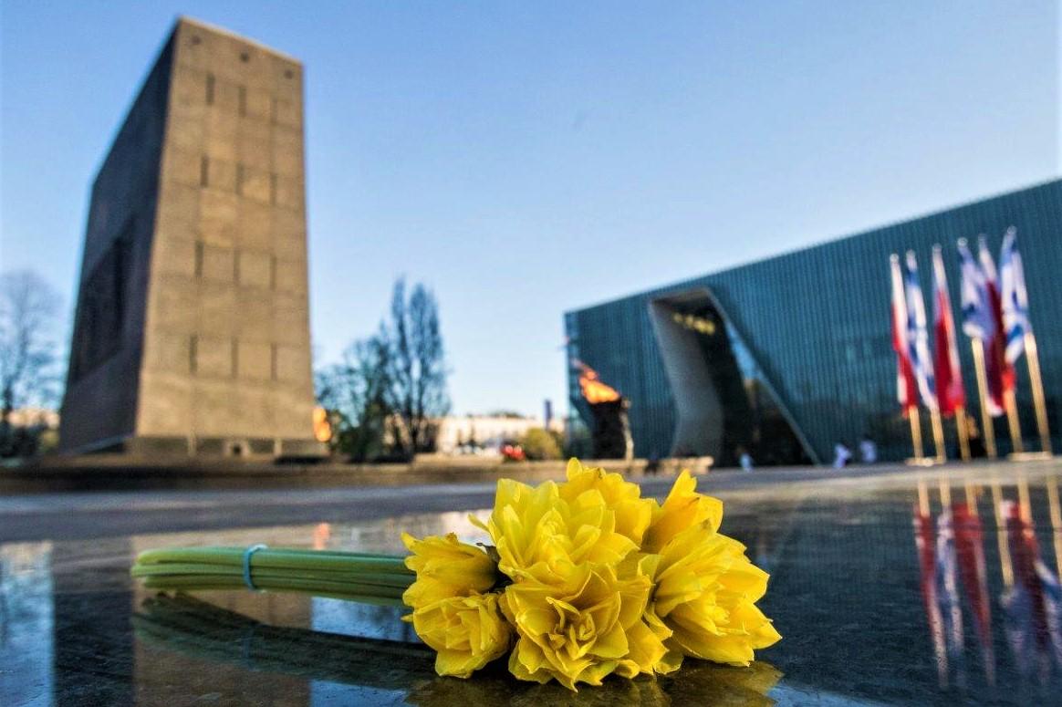 Mały bukiet żonkili leży na płycie nieopodal pomnika Bohaterów Getta. W tle budynek Muzeum POLIN i flagi polsko-izraelskie.