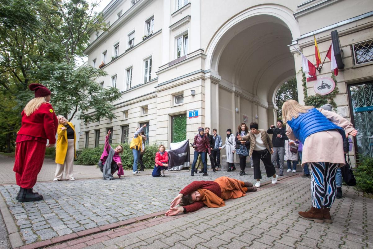 Grupa osób stoi w półkole przy jednej z kamienic na warszawskim Muranowie. Pomiędzy tymi osobami na chodniku leży kobieta.