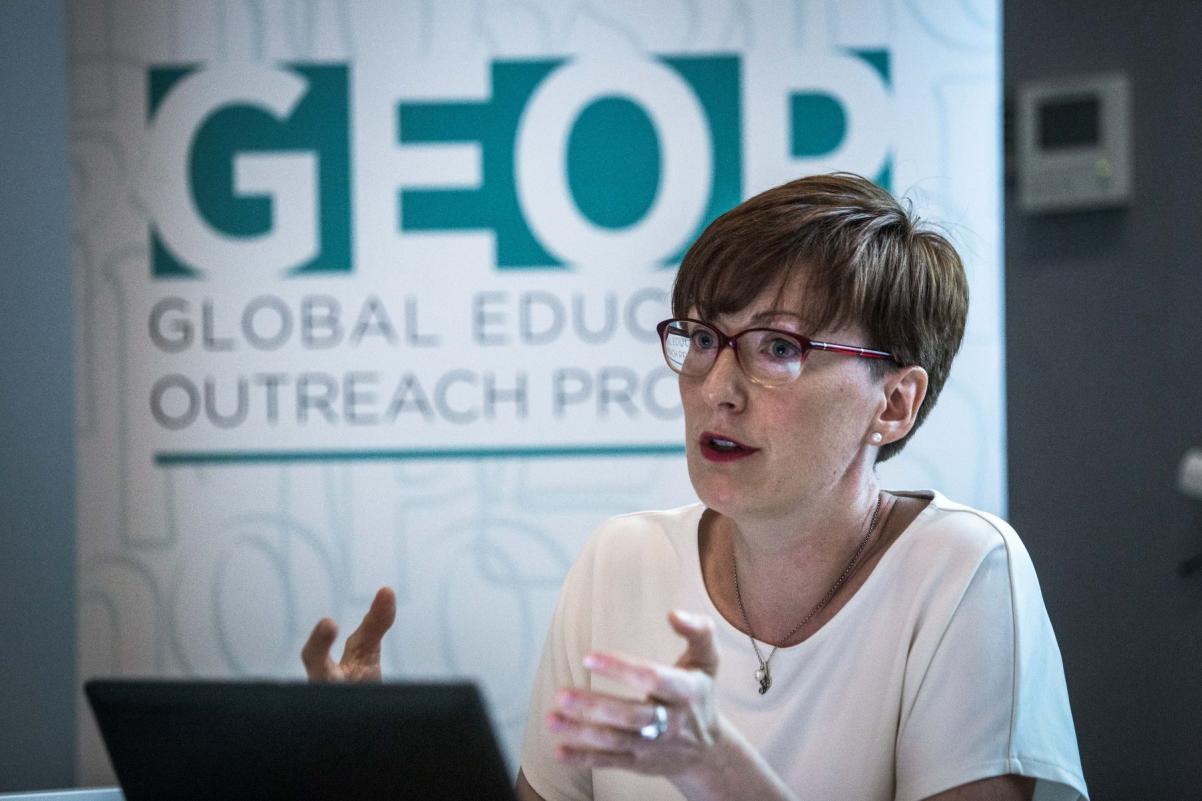 Kobieta w okularach tłumaczy coś podczas konferencji. W tle baner z logo Global Education Outreach Program.
