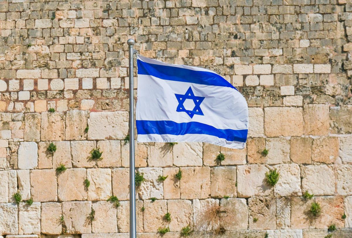 Ściana z kamiennych bloków, poprzerastana roślinnością. Na dole kamienne bloki są dużo większe niż na górze. Przed murem flaga Izraela, biała, z granatowymi pasami biegnącymi wzdłuż górnej i dolnej krawędzi. Na środku granatowa gwiazda Dawida.