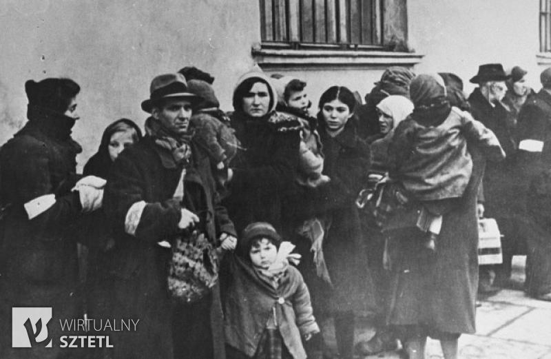Przy jakimś budynku stoją Żydzi z getta krakowskiego zgromadzeni przed deportacją. Zdjęcie czarno-białe. EN: The Jews from Krakow Ghetto before deportation. They are standing next to some building. A photo is black and white.