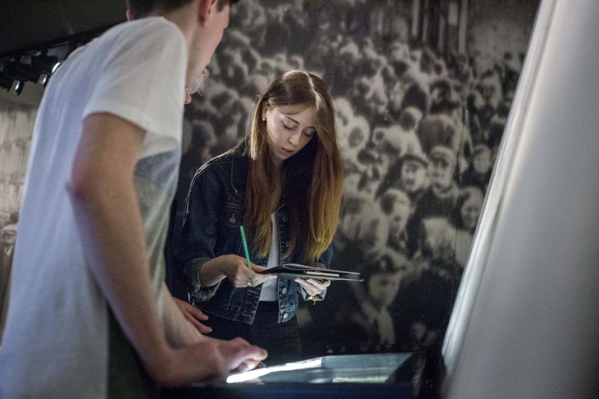 Dziewczyna i chłopak stoją przy gablocie na wystawie stałej w Muzeum POLIN. Dziewczyna trzyma notes i długopis.