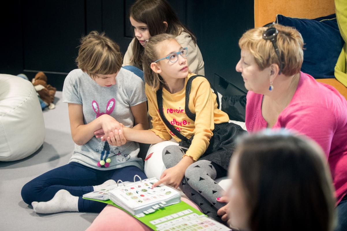 Na podłodze siedzą trzy dziewczynki. Jedna z nich wskazuje obrazek narysowany w książce. Obok niej siedzi kobieta.