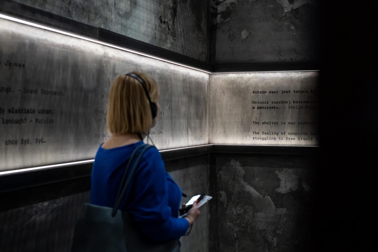 Kobieta stoi w bunkrze zrekonstruowanym na wystawie "Wokół nas morze ognia". Zwiedzająca ma słuchawki na uszach, a przed sobą trzyma audioprzewodnik. Na ścianach bunkra znajdują się zapiski ze wspomnień osób ukrywających się w getcie warszawskim.