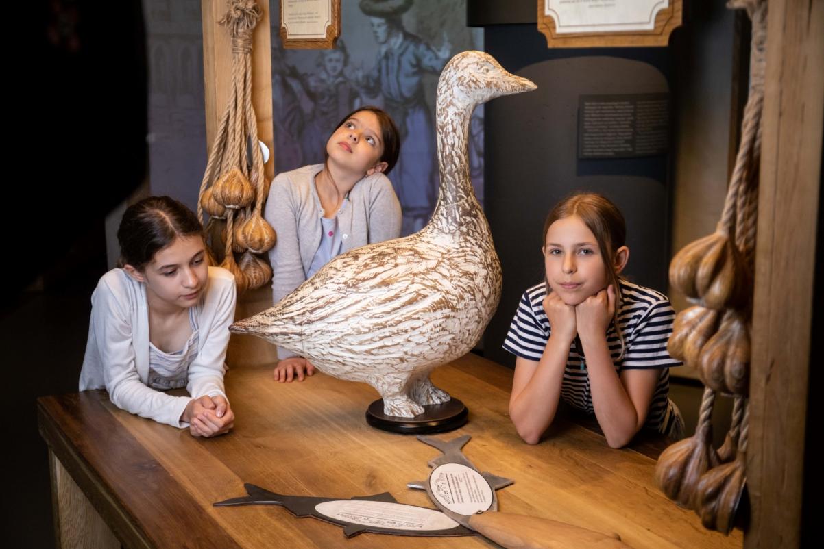Trzy dziewczynki opierają się o stół na wystawie stałej w Muzeum POLIN, na którym stoi figurka gęsi, a z góry zwisa sztuczny czosnek.