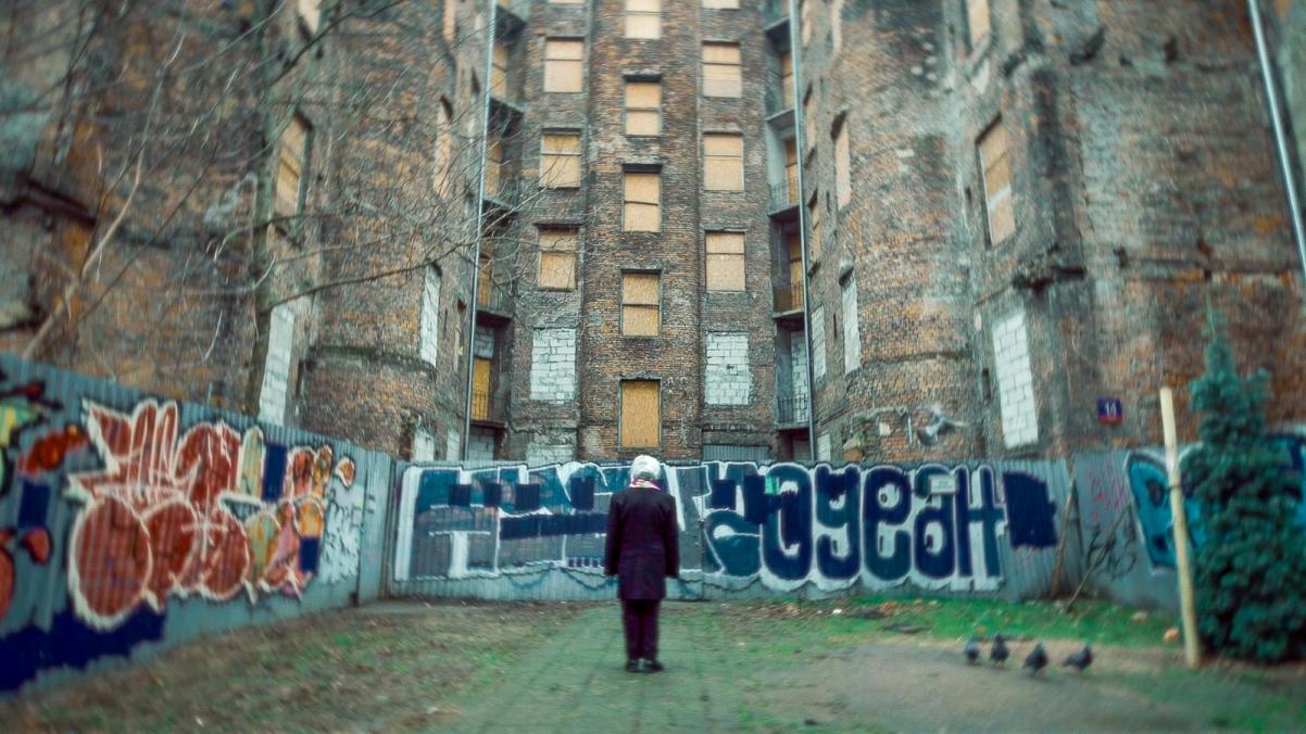 Starsza kobieta stoi przed starą zniszczoną kamienicą, ogrodzoną blachą z graffiti.