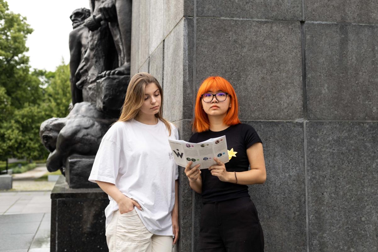 Dwie dziewczyny stoją oparte o pomnik Bohaterów Getta. Dziewczyna z pomarańczowymi włosami trzyma przed sobą ulotkę, na którą spogląda jej brązowowłosa koleżanka.