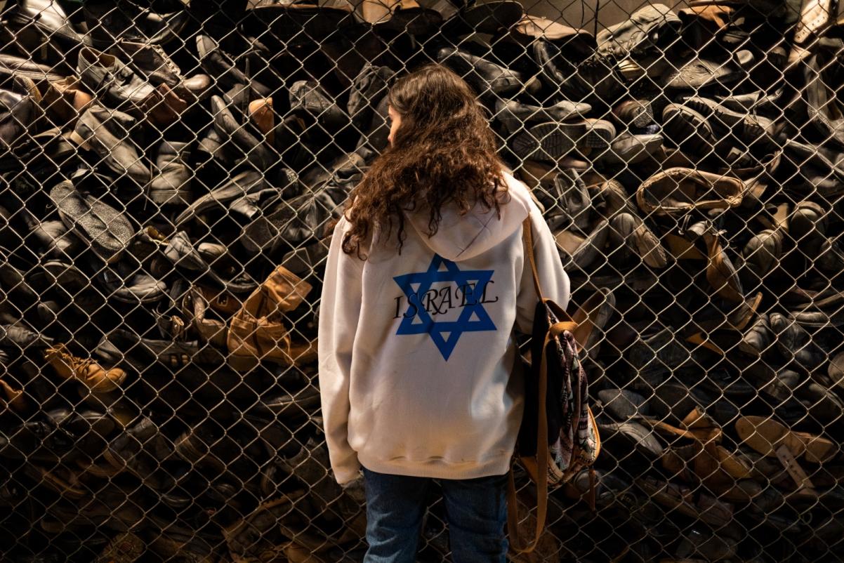 Muzeum Auschwitz-Birkenau. Dziewczyna w bluzie z gwiazdą Dawida stoi przed ścianą z butami więźniów obozu Zagłady w Auschwitz.