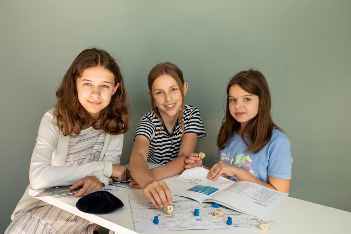 Trzy uśmiechnięte dziewczyny siedzą przy stole i bawią się zestawem edukacyjnym "Muzeum w pudełku. Miasteczko Malki".