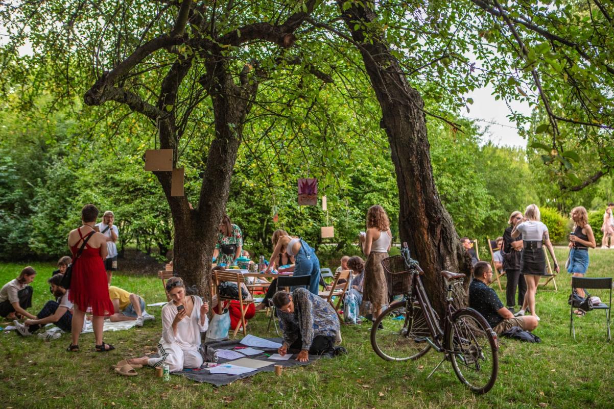 Ludzie stoją lub siedzą na trawie pod drzewami w parku.