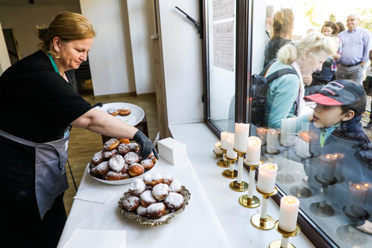 Kobieta nakłada pączki na talerz. Dziecko czeka na wypieki, stojąc za szybą.