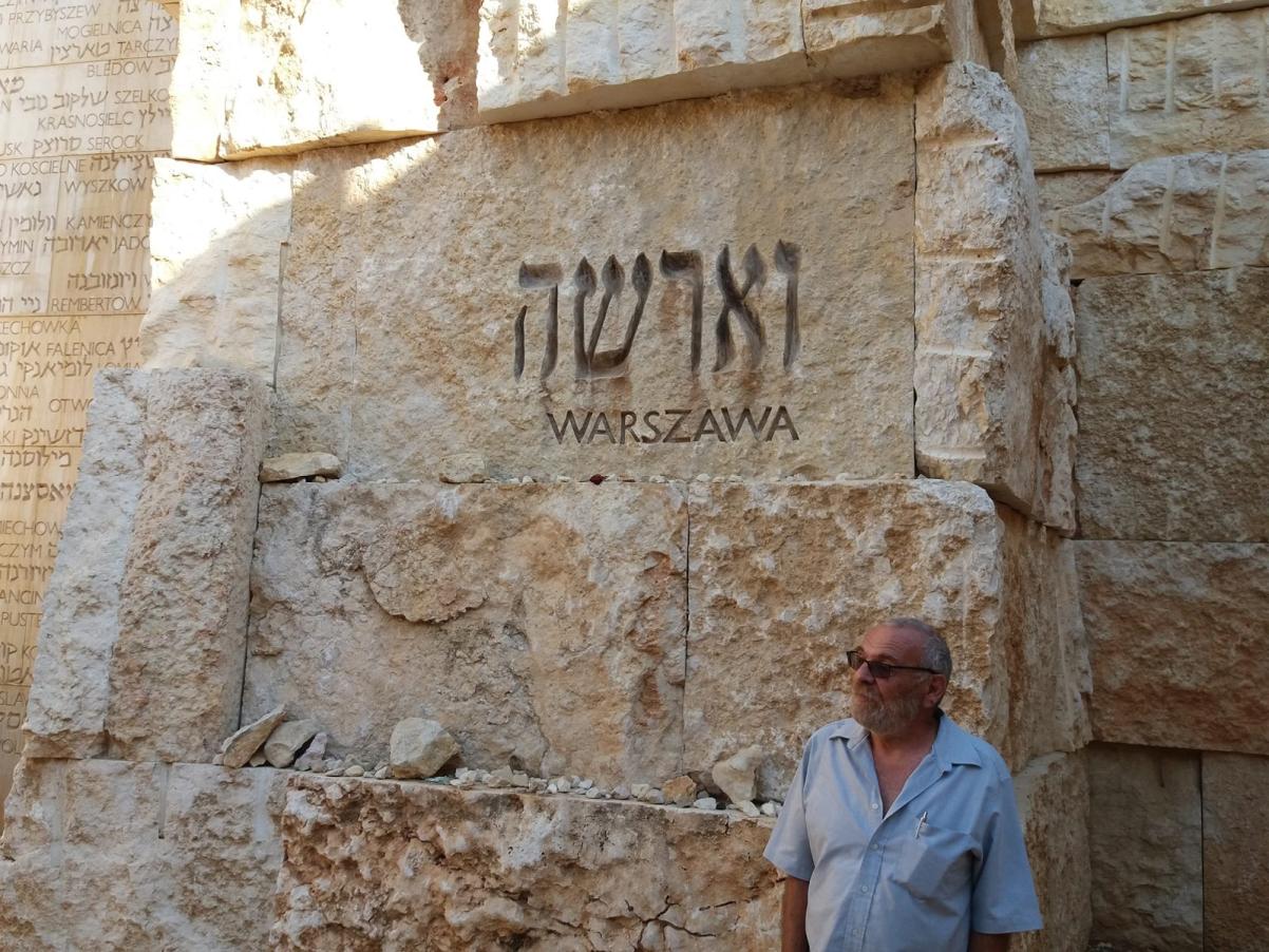 Mur z kamieni w kolorze piasku, z napisami "Warszawa, w języku hebrajskim i polskim. W prawym dolnym roku mężczyzna - Alex Dancyg - w jasnej letniej koszuli.