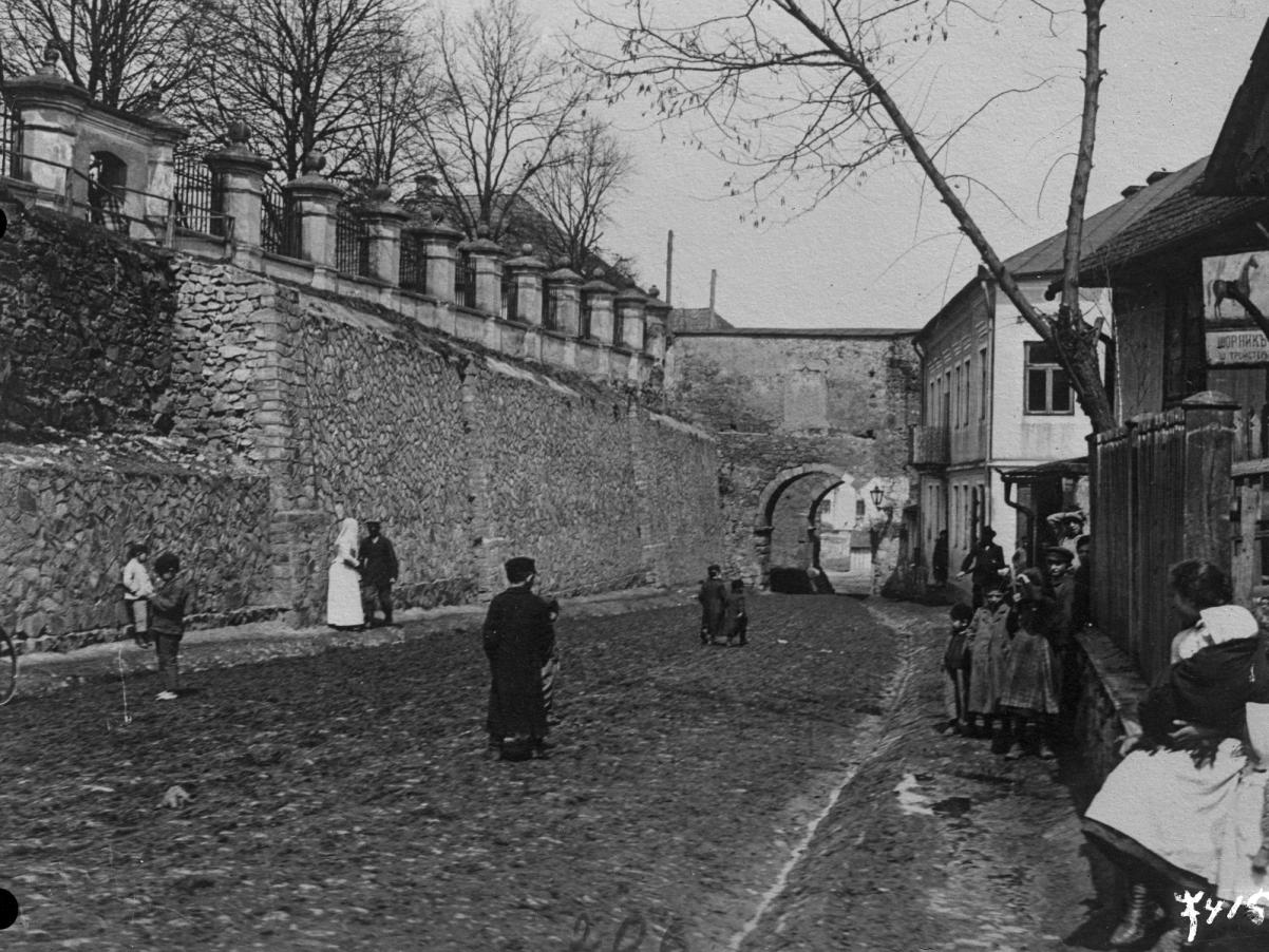 Po brukowanej ulicy jakiegoś miasteczka spaceruje kilka osób. English description: A few people are walking along the cobbled street of a town.